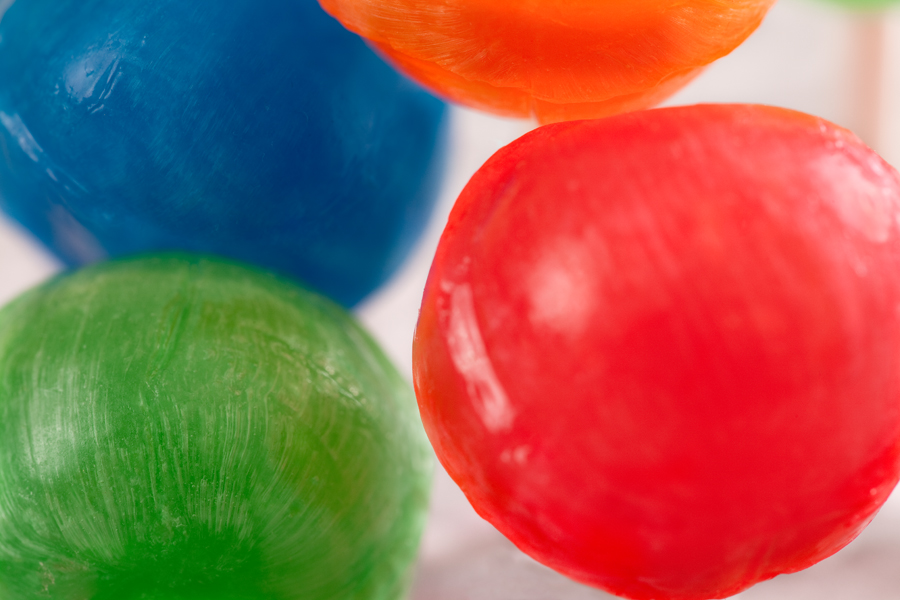 This Brian Charles Steel image is composed of the tops of four lollipops in front of a light-grey background. The lollipops occupy most of the image. The closest lollipop is red, and it takes up ninety-percent of the right half of the frame. Part of an orange one takes up a large portion of the remaining ten-percent of the right side. The bottom left half of the frame is filled with a green lollipop, and behind it, filling the top-half, is a blue lollipop. 